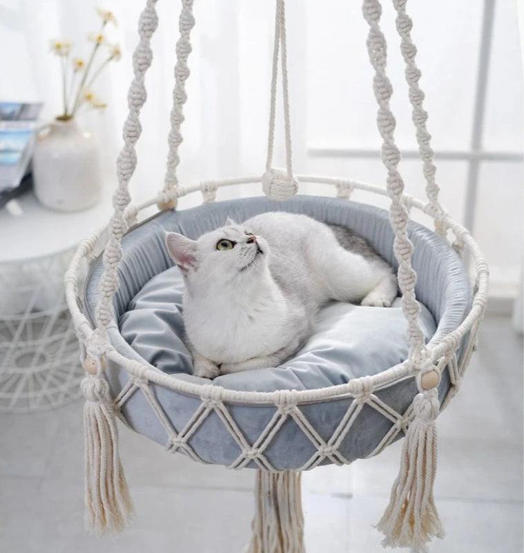 White cat resting on a cushioned macrame cat swing bed, providing comfort and style in a bright indoor setting.