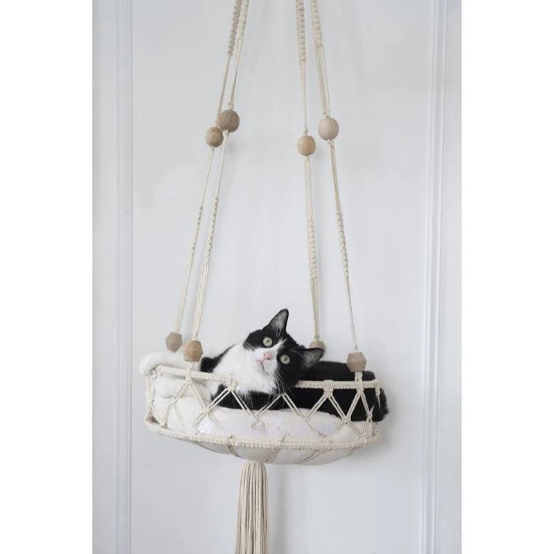 Black and white cat lounging in a macrame cat swing bed, suspended with decorative wooden beads and tassels.